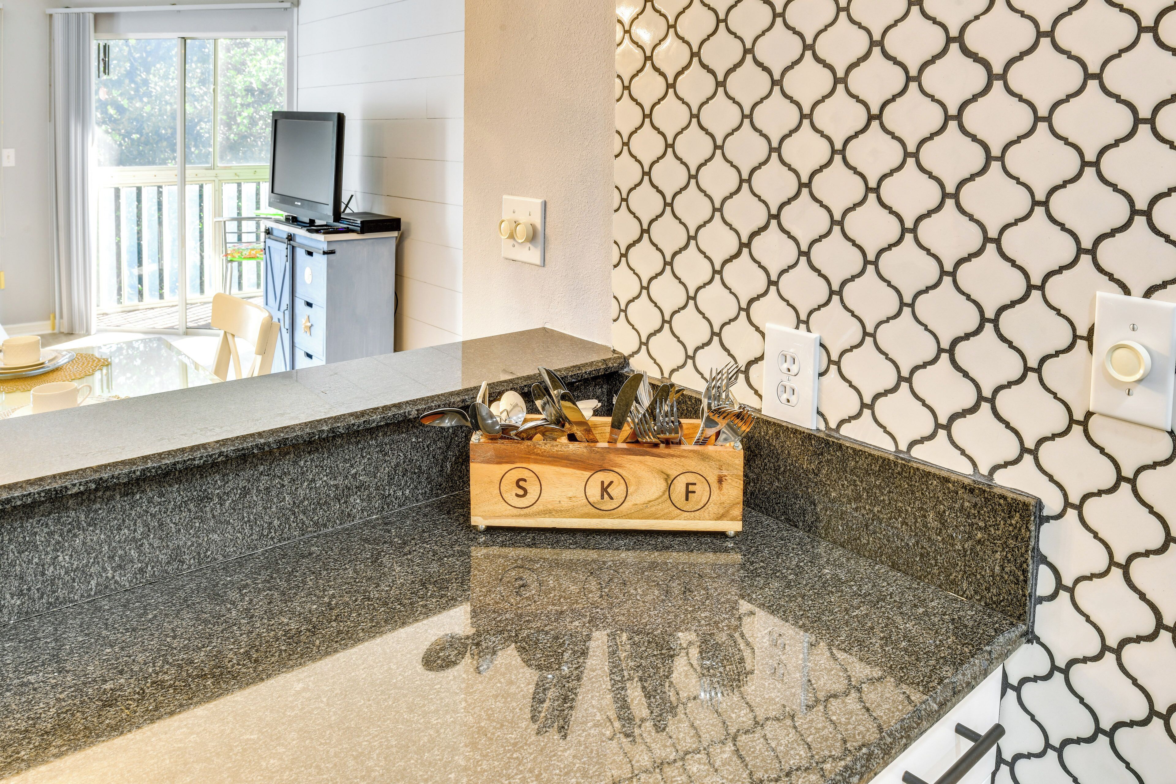 A kitchen with a granite countertop, a wooden utensil holder, a television on a stand, and a patterned backsplash.