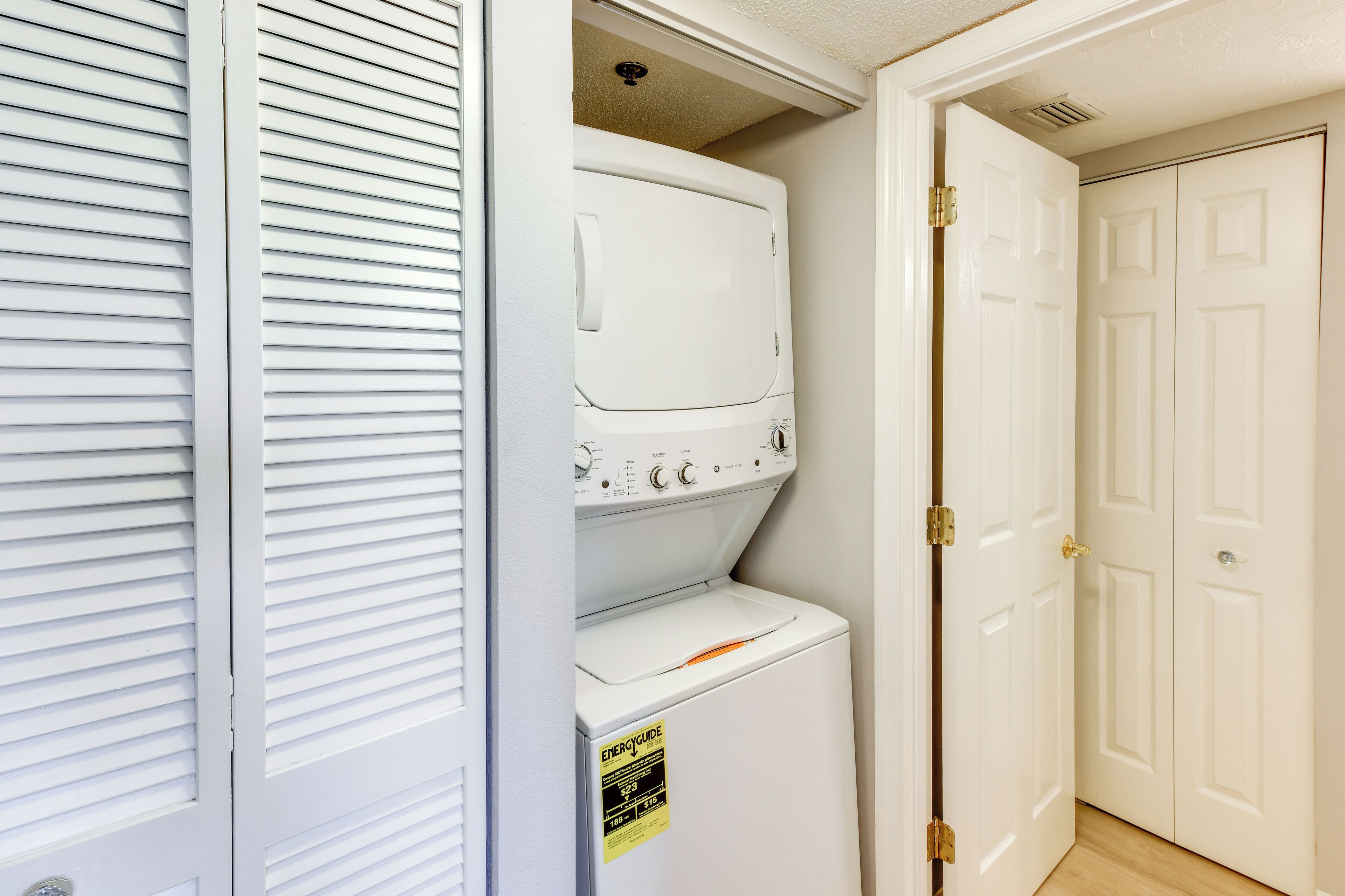 A laundry room with a stacked washer and dryer, a door with a brass handle, and a white wall with a vent.