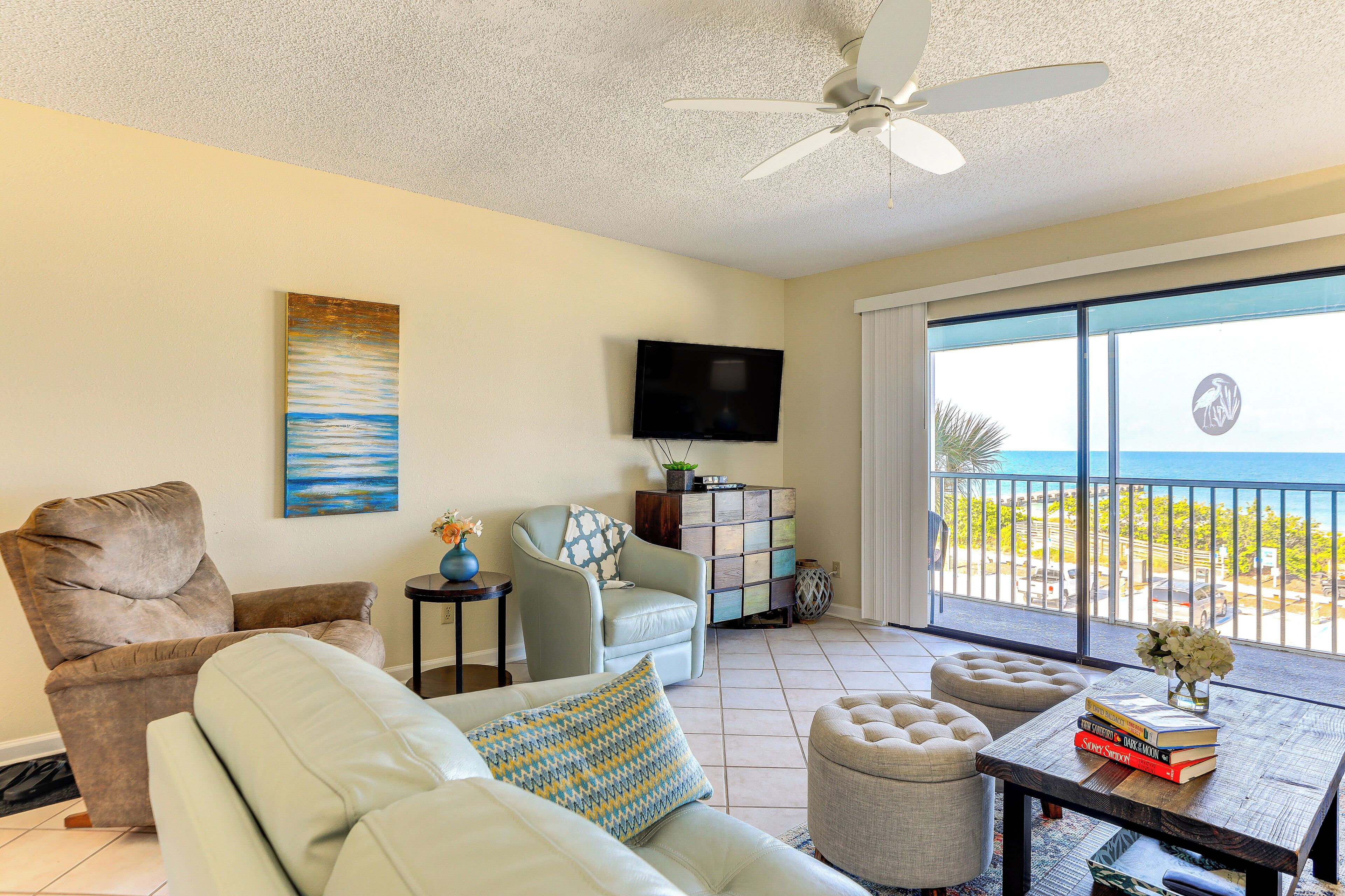 A living room with a sofa, armchair, and ottoman, a coffee table with books, a TV mounted on the wall, and a balcony with a view of the ocean.