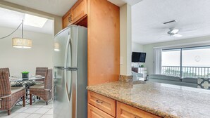 A kitchen with a granite countertop, wooden cabinets, a stainless steel refrigerator, wicker chairs, a dining table, and a large window with a balcony. - Pelican Cove Condo at Bradenton Beach w/ Pool! (Bradenton Beach)