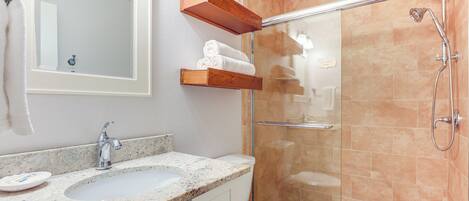 Un baño con lavamanos de encimera de granito, inodoro blanco, cabina de ducha de vidrio y estantes de madera sobre el inodoro.