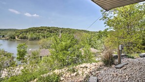 A lakeside view with a covered outdoor area, rocky terrain, and a distant forested hill. - Lake Views, Fire Pit! Blue Cabin Ozarks Getaway (Camdenton)