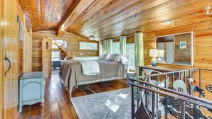 A log cabin bedroom with a bed, a small table, a mirror, and a window with curtains.