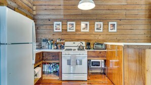 A compact kitchen with wooden walls, a white refrigerator, a stove, an oven, and a microwave. There are framed pictures on the wall and a hanging light fixture.