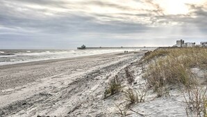 A beach with a pier extending into the ocean, sandy dunes, and buildings in the background.