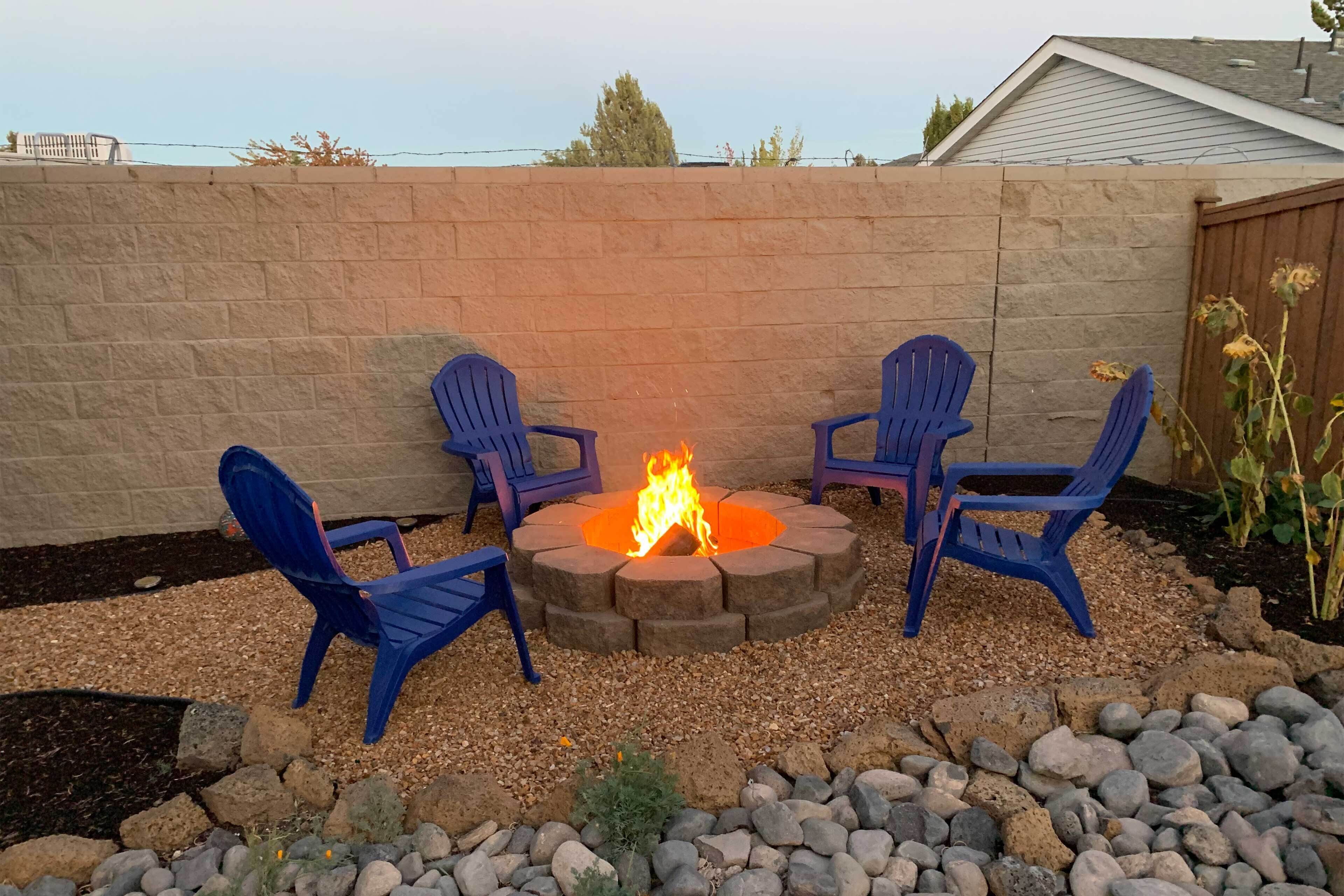 A fire pit with four blue chairs arranged around it, set against a stone wall with a garden area in front.