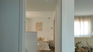 A narrow hallway leading to a bedroom with a bed and a small table, a radiator, and a window with curtains. - Atico-loft Mompó. Lujo Marítimo (València)