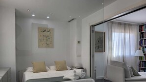 A modern bedroom with a bed, bedside table, and a chair. There is a bookshelf with books and a painting on the wall. - Atico-loft Mompó. Lujo Marítimo (València)