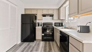 A compact kitchen with a black refrigerator, stove, and dishwasher, beige cabinets, and a white countertop.