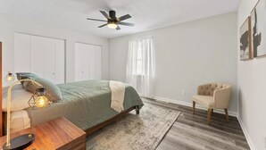 A bedroom with a bed, a chair, a desk, a lamp, a ceiling fan, and a window with curtains.