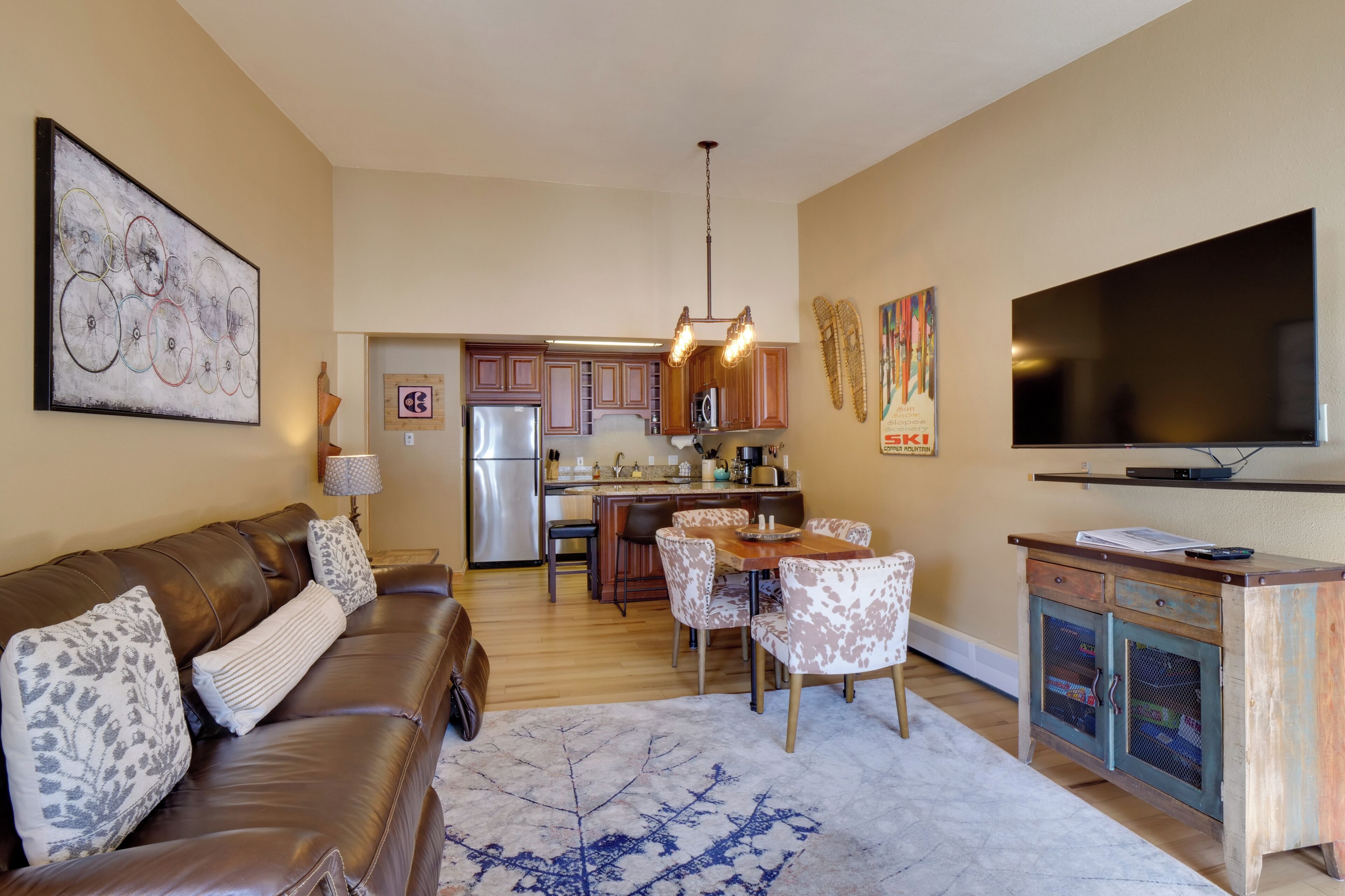 A modern living room with a brown leather sofa, a dining table with patterned chairs, a large flat-screen TV, and a kitchen in the background.