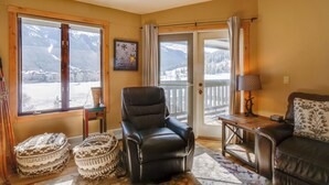 Un salón acogedor con un sillón de cuero, una mesa de madera y un sofá. Amplias ventanas ofrecen vistas a montañas cubiertas de nieve.