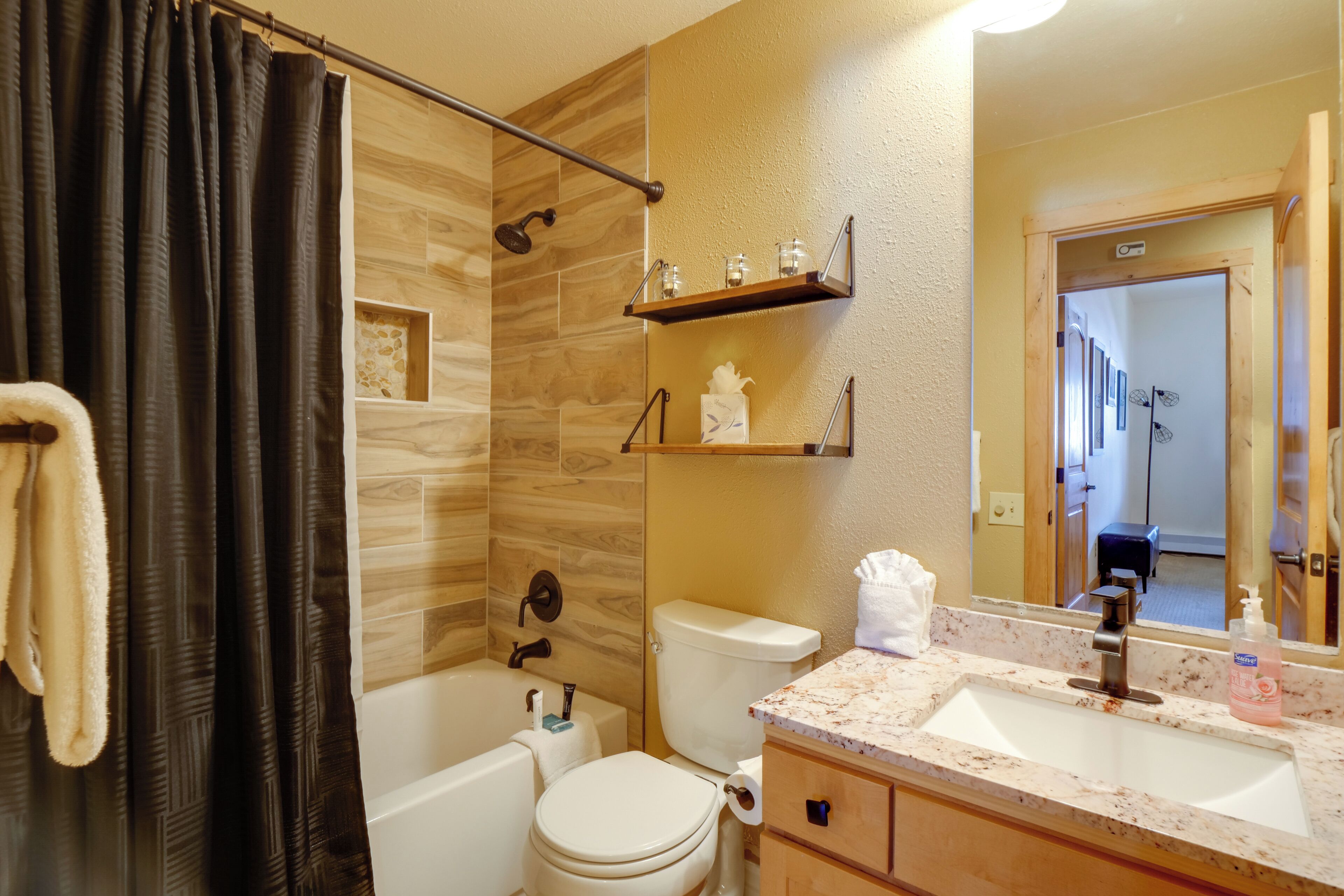 A bathroom with a bathtub, toilet, and sink. There are shelves above the sink and a shower curtain.