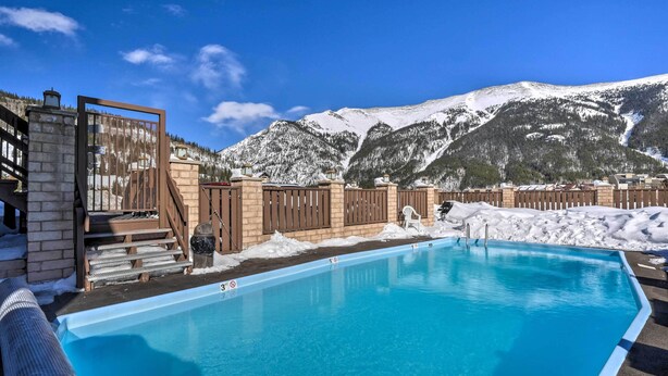 Una piscina con montañas al fondo, muelle de madera y pared de piedra.