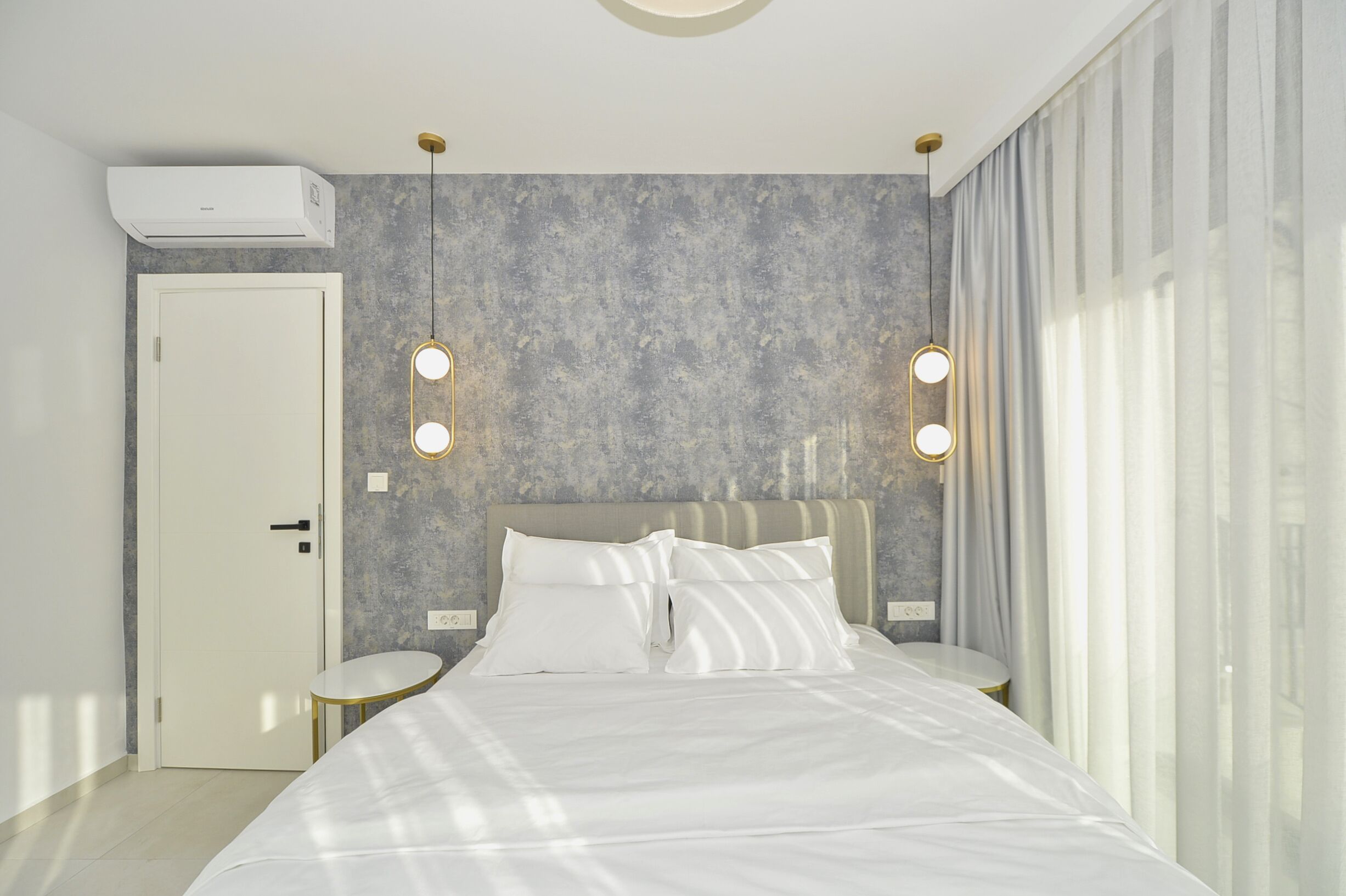 A modern hotel room with a large bed, two bedside tables, a wall-mounted air conditioner, and a large window with sheer curtains.