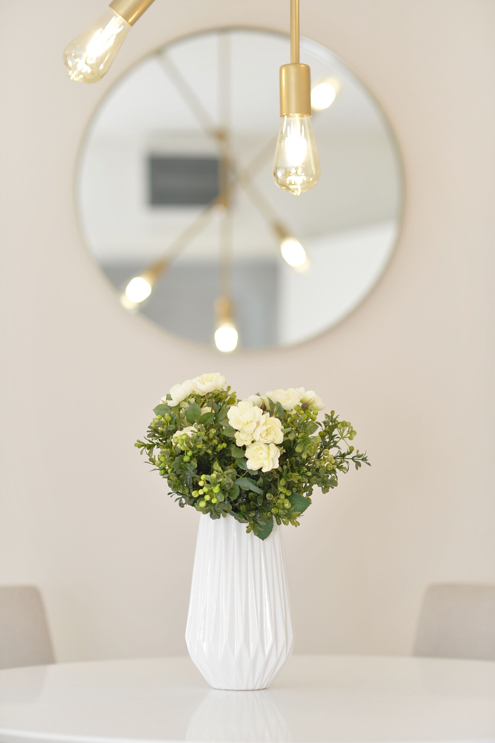 Un jarrón blanco con flores sobre una mesa, un espejo redondo e iluminación colgante.