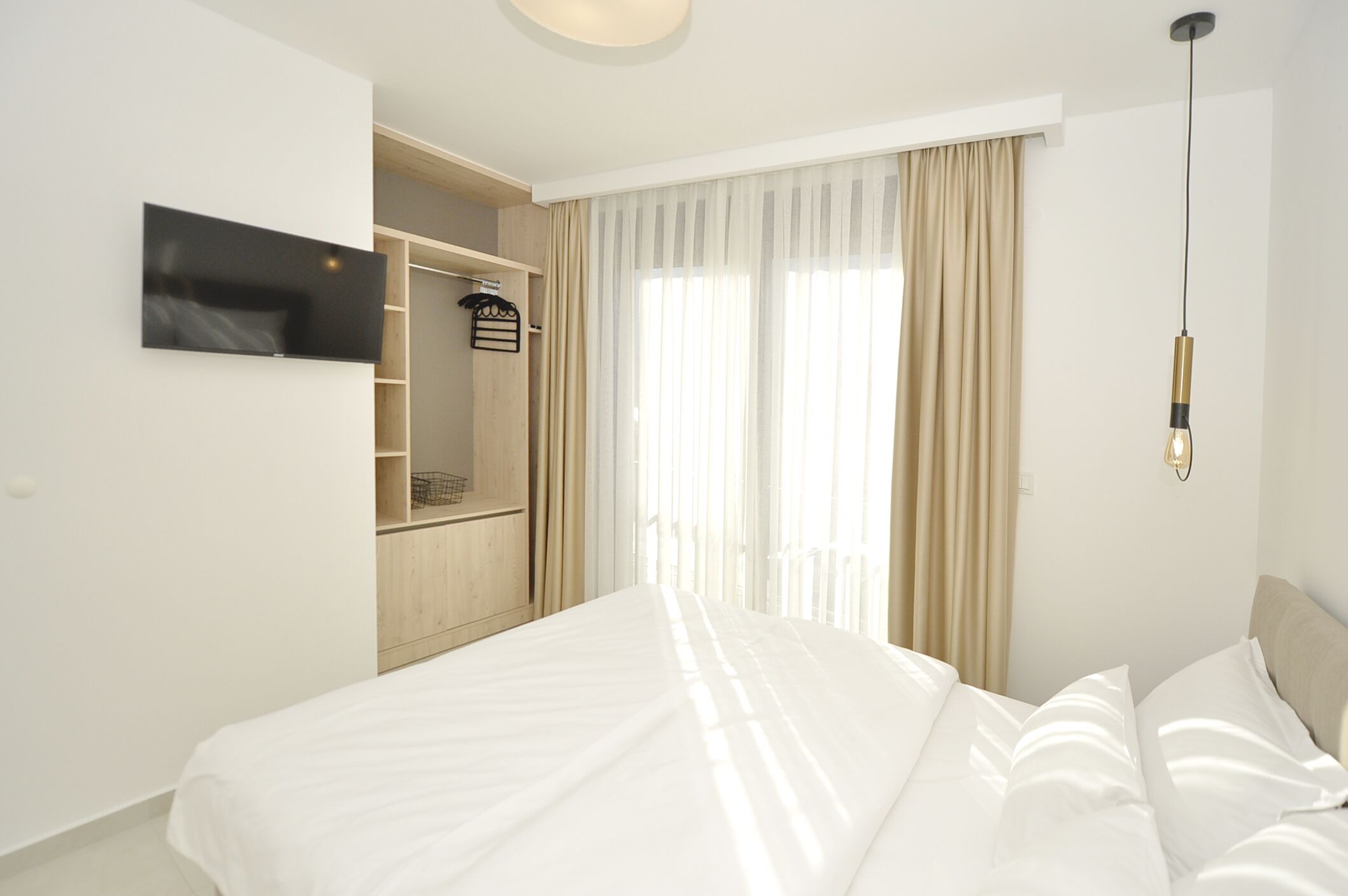 A modern hotel room with a large bed, a TV mounted on the wall, a wardrobe, and a window with sheer curtains.