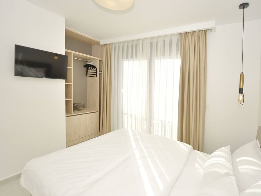 Una habitación de hotel moderna con una cama grande, un televisor en la pared, un armario y una ventana con cortinas de gasa.