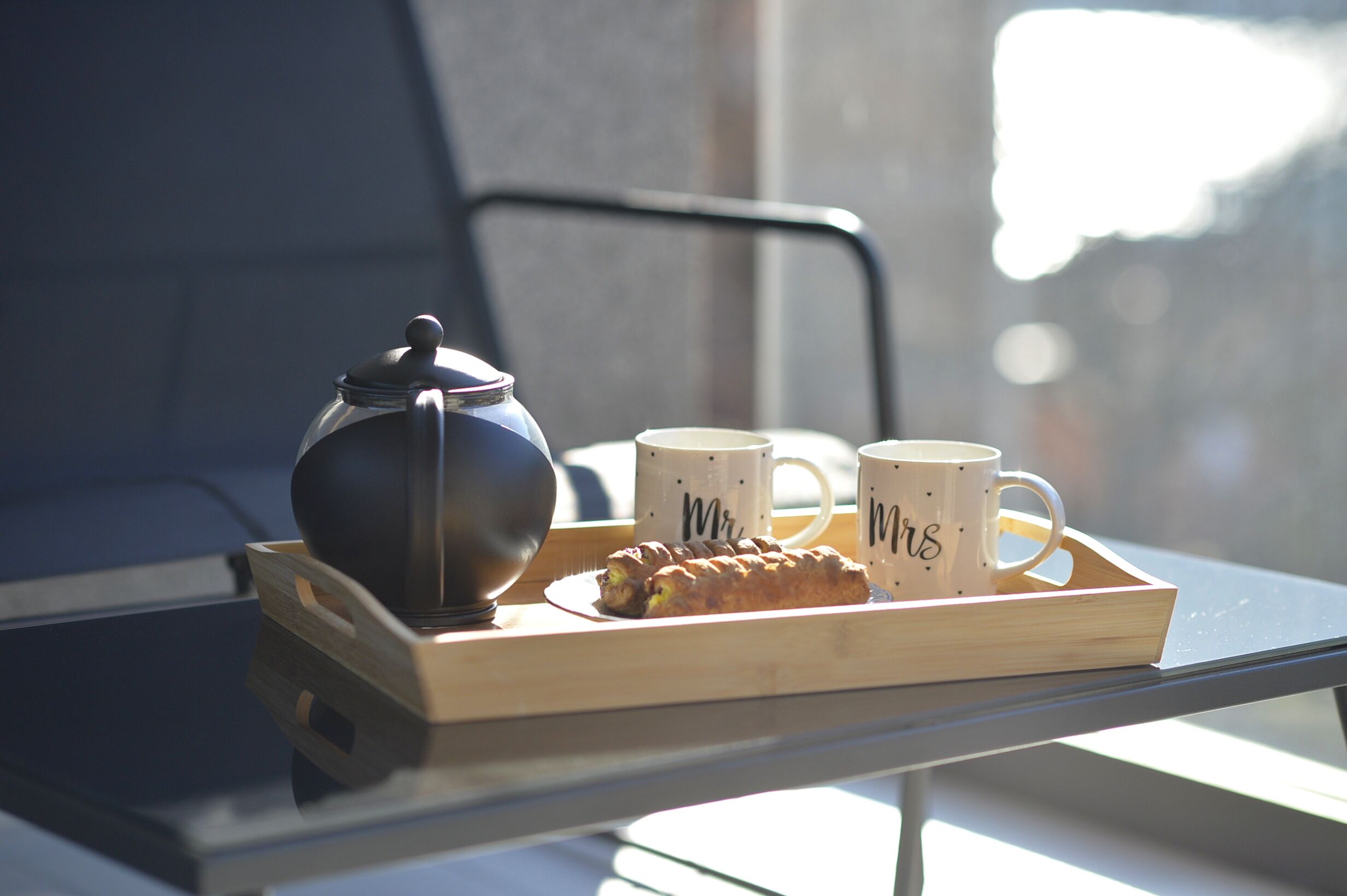 Een dienblad met een theepot, twee mokken en een gebakje op een tafel bij een raam.