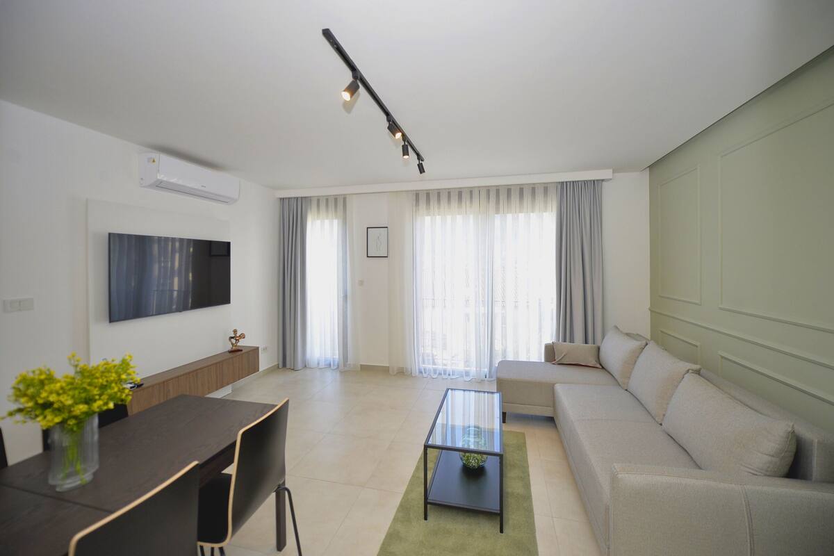 Una sala de estar moderna con un sofá, una mesa de centro de vidrio, un televisor montado en la pared y una zona de comedor con una mesa y sillas.