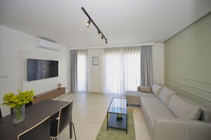 Una sala de estar moderna con un sofá, una mesa de centro de vidrio, un televisor montado en la pared y una zona de comedor con una mesa y sillas.