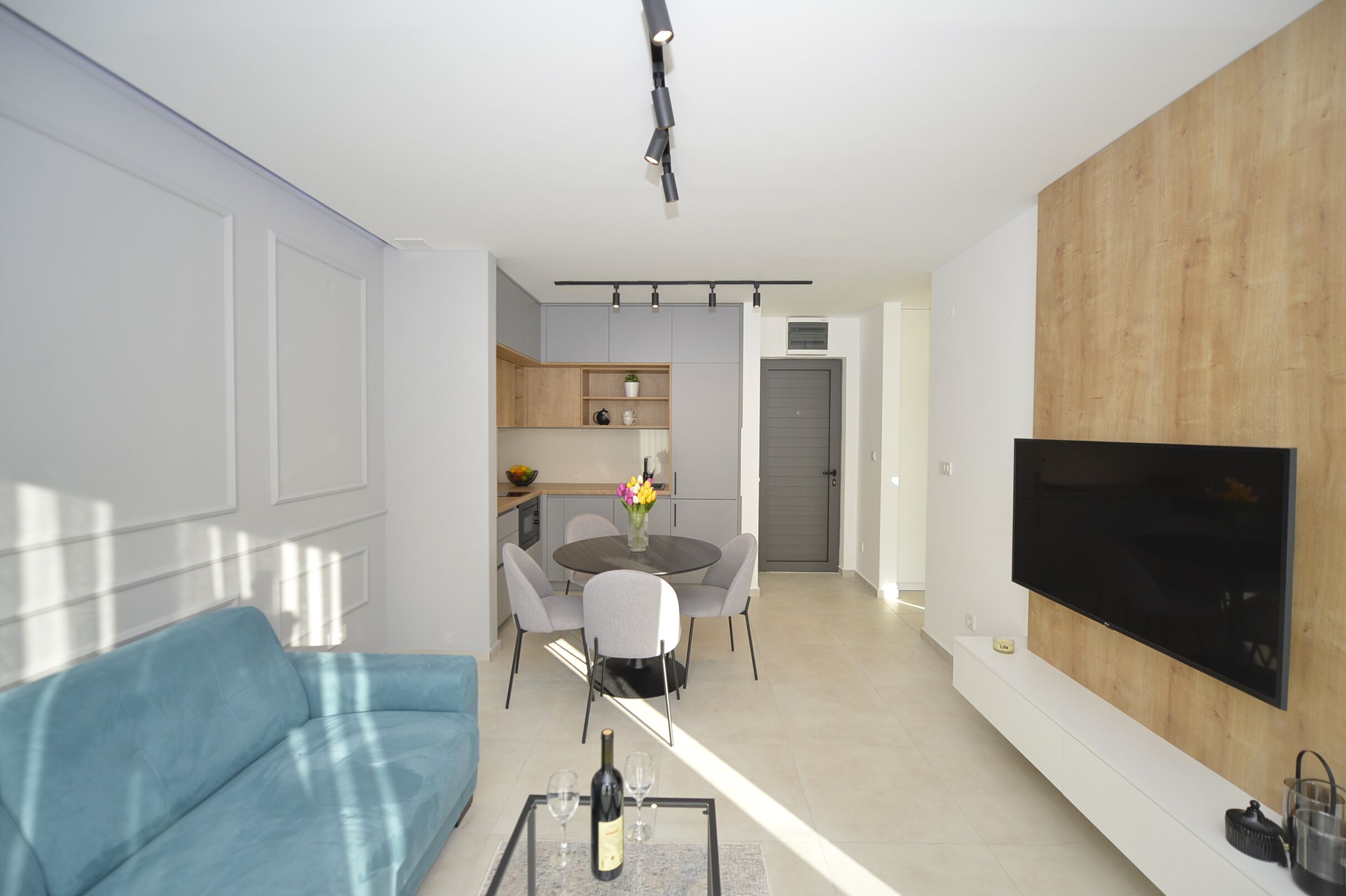 Uma sala de estar moderna com um sofá azul, um espaço de jantar com uma mesa redonda e cadeiras, uma cozinha com armários de madeira e uma TV na parede.
