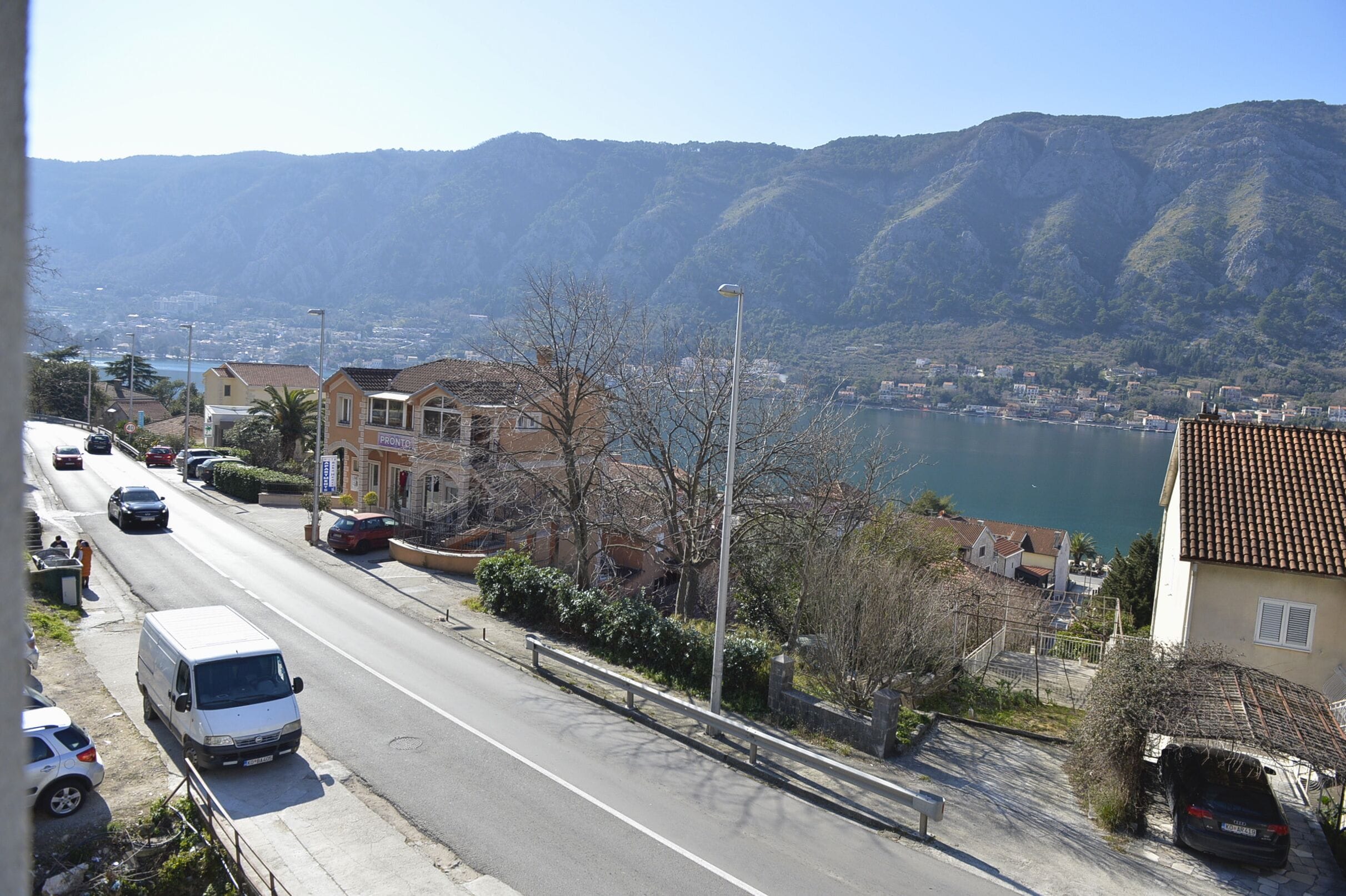 Una carretera costera con edificios residenciales, una camioneta blanca y vistas al mar y a las montañas.