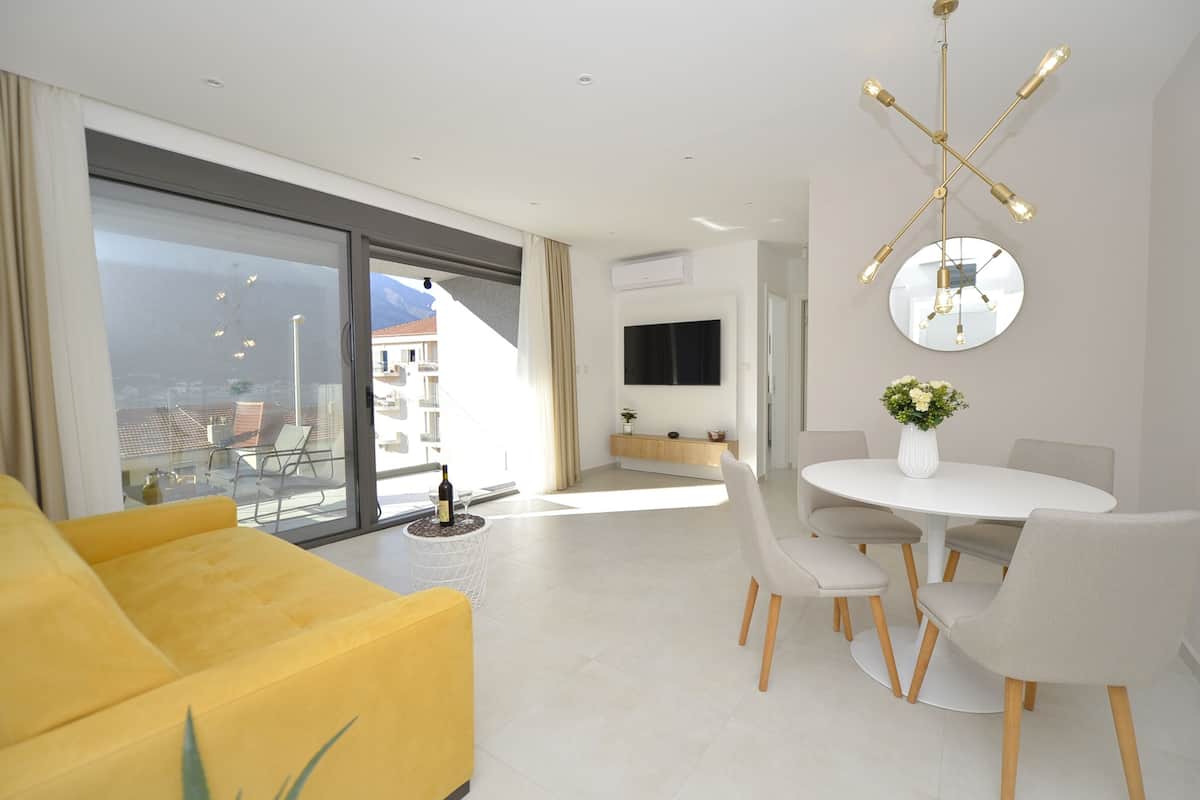 Una sala de estar moderna con un sofá amarillo, una mesa de comedor con sillas, un televisor montado en la pared y un ventanal con vistas a edificios.