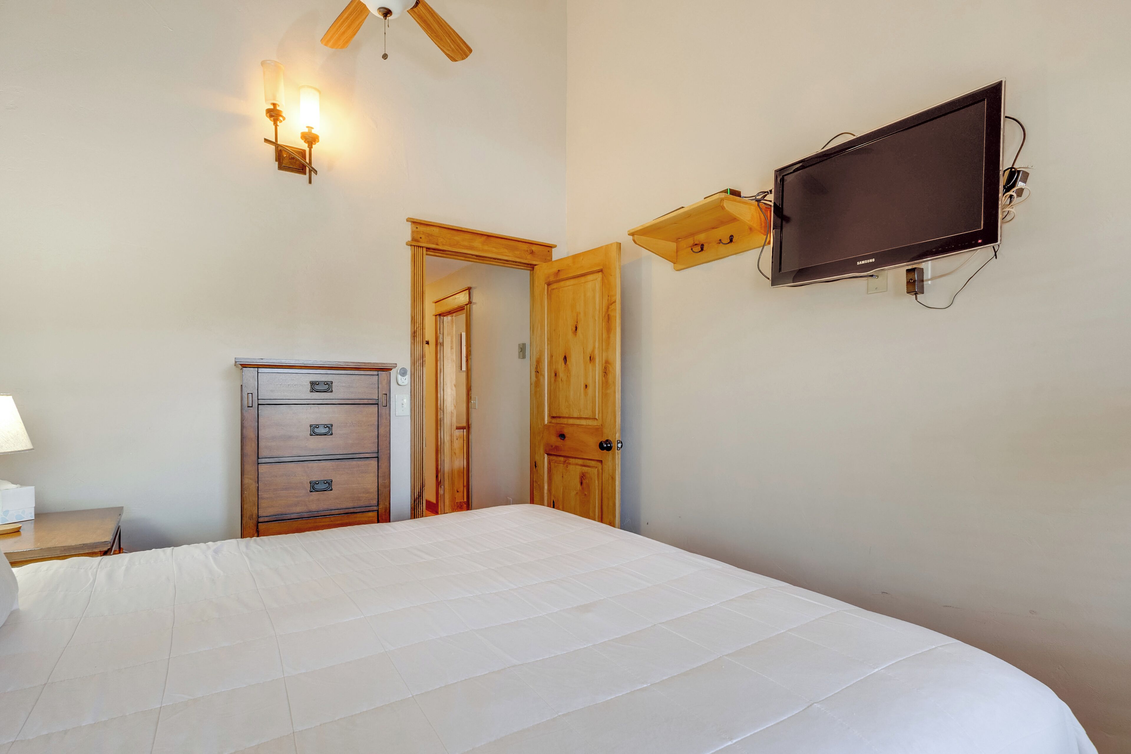 A bedroom with a bed, a wooden dresser, a television mounted on the wall, and a ceiling fan.