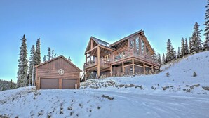 A wooden cabin with a garage and a large deck, surrounded by snow and trees.
