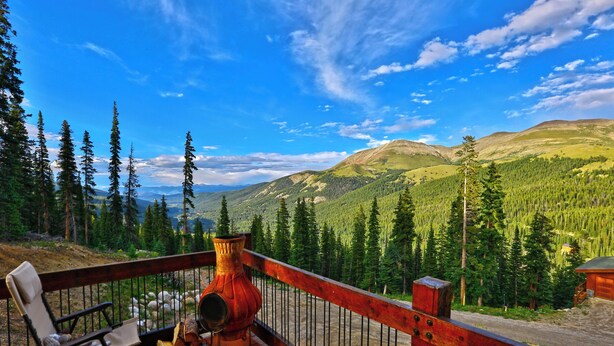 A wooden deck with chairs and a rustic vase, overlooking a mountainous landscape with dense forests and a clear blue sky.