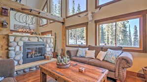 A cozy cabin interior with a stone fireplace, wooden furniture, and large windows overlooking a forest.