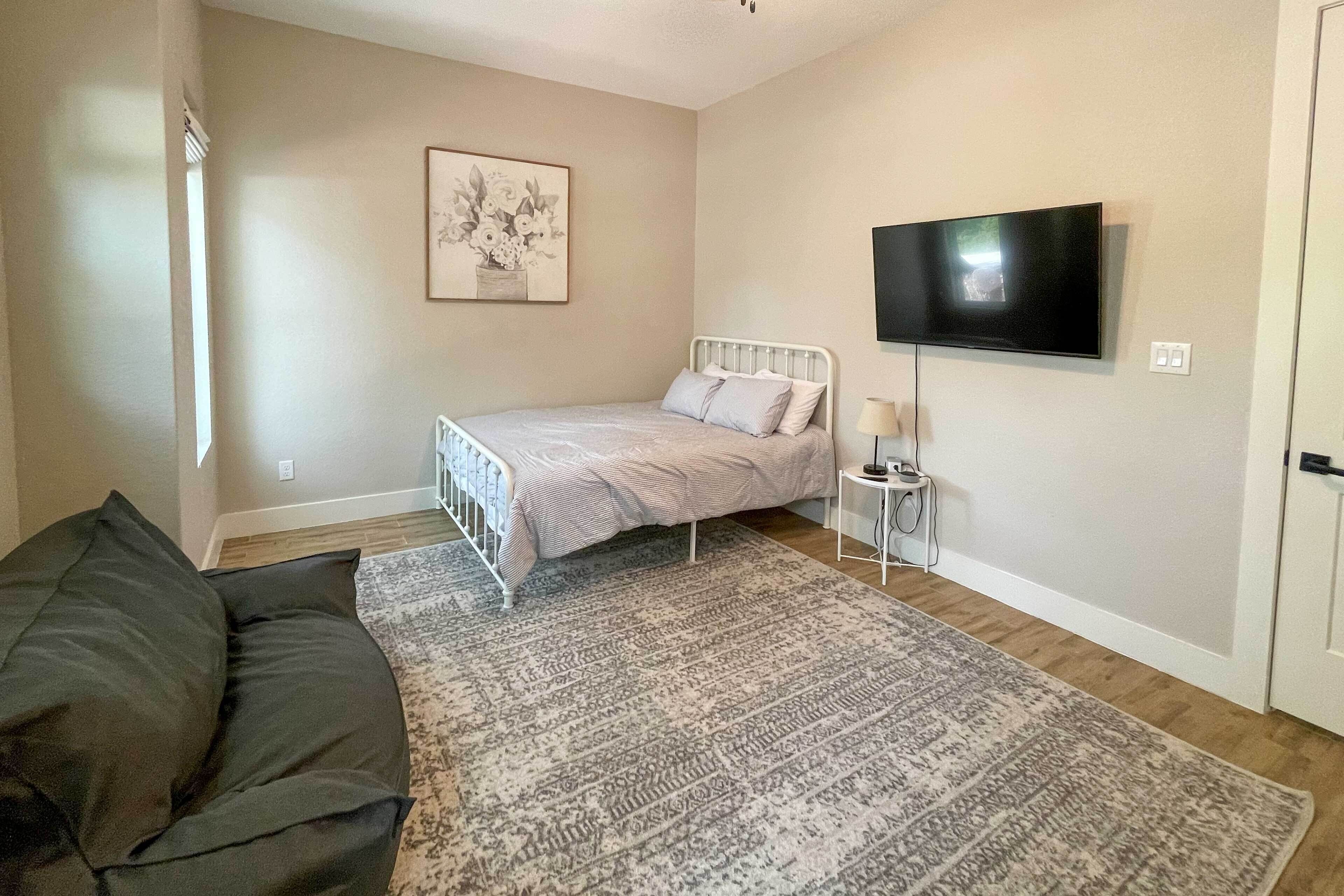 Un dormitorio con una cama, televisión, mesita de noche, lámpara de pie, alfombra estampada y un sofá.