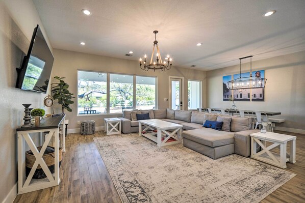 A spacious living room with a large sectional sofa, a coffee table, and a chandelier. There are large windows providing natural light and a view of the outdoors. - Brand New Modern Canyon Lake Escape w/ Fire Pit! (Canyon Lake)