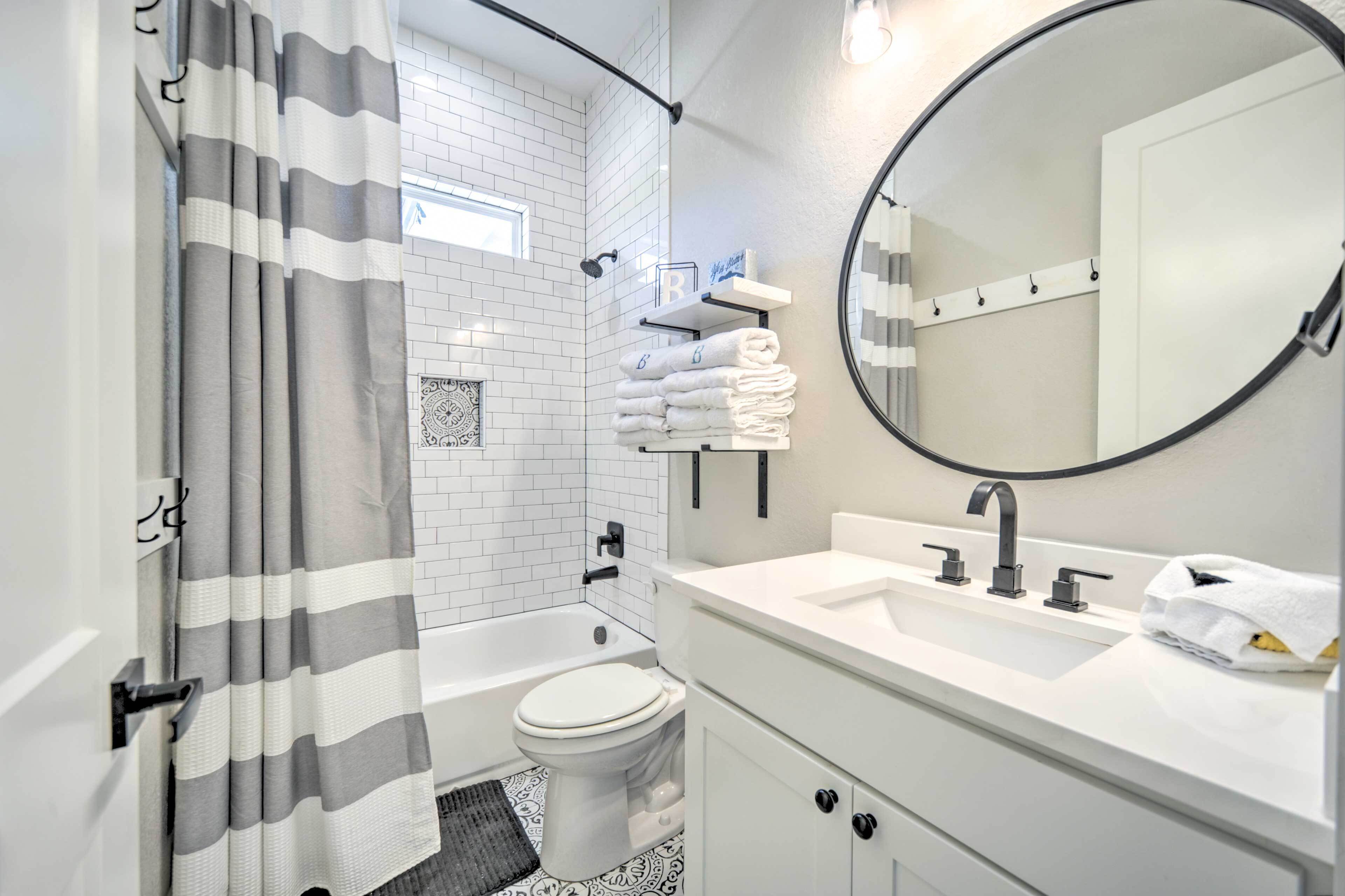 A modern bathroom with a bathtub, toilet, and a large mirror. There are towels stacked on a shelf and a shower curtain.