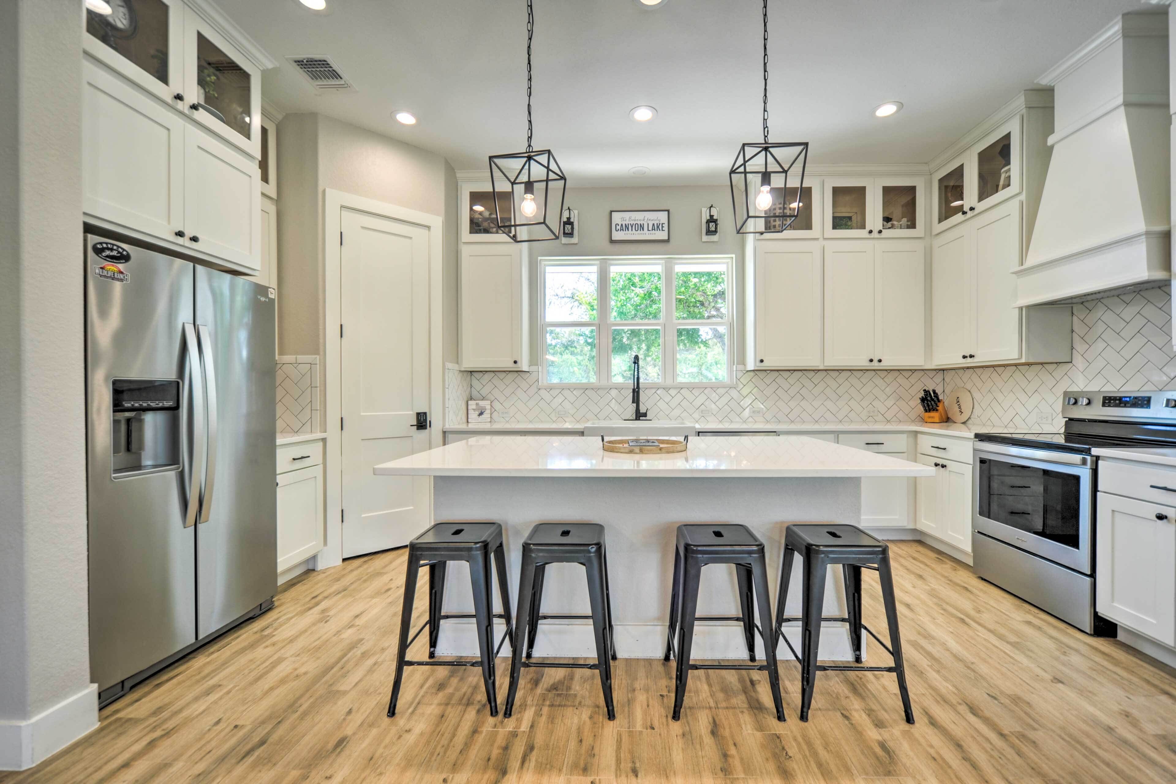A modern kitchen with white cabinetry, a stainless steel refrigerator, a central island with stools, and a window with a view of greenery.
