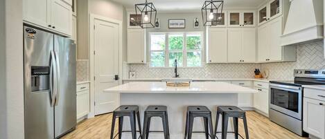 A modern kitchen with white cabinetry, a stainless steel refrigerator, a central island with stools, and a window with a view of greenery.