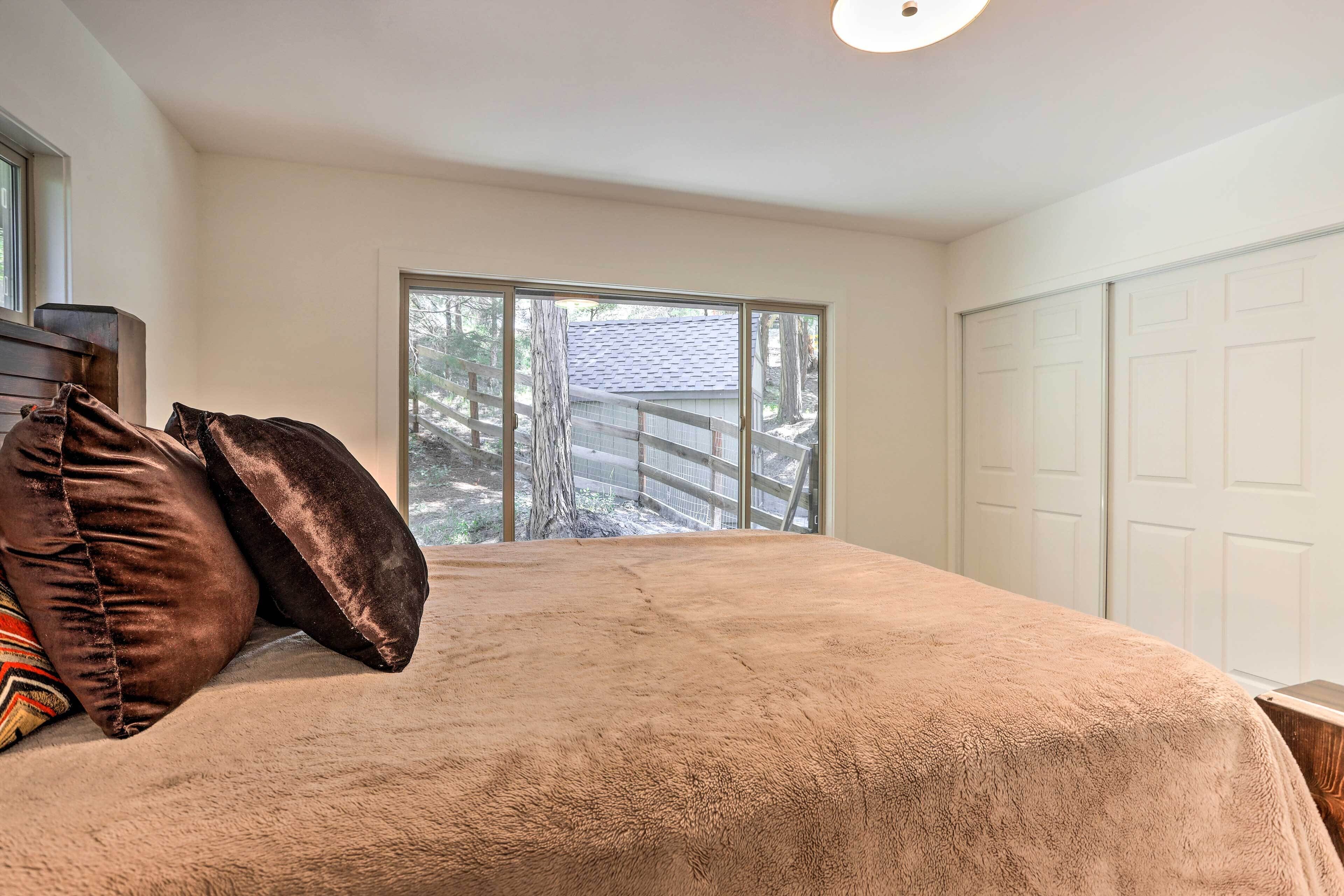 Un dormitorio con un ventanal grande, una cama con manta color beige y cabecero de madera.