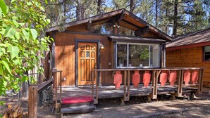 A rustic cabin with a wooden door, a porch with red railings, and a window overlooking a forested area.