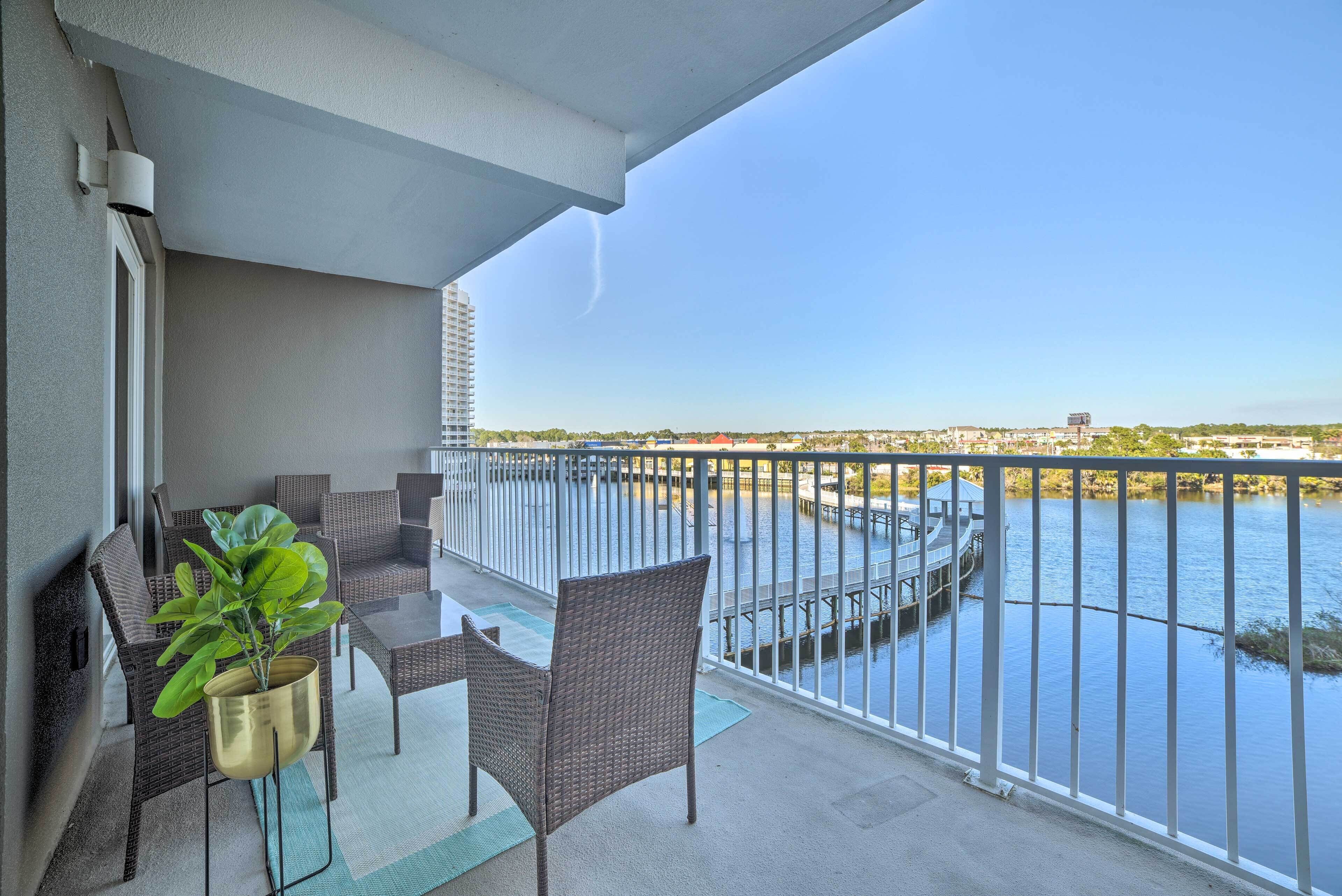 A balcony with wicker furniture, a potted plant, and a view of a body of water with a dock and buildings in the distance.