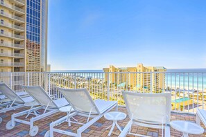 A balcony with white lounge chairs and a small table, overlooking a beach and hotel buildings.