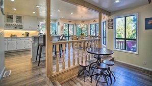 Una cocina amplia con armarios blancos, una zona de comedor con barandal de madera y ventanas grandes que ofrecen vistas al exterior.