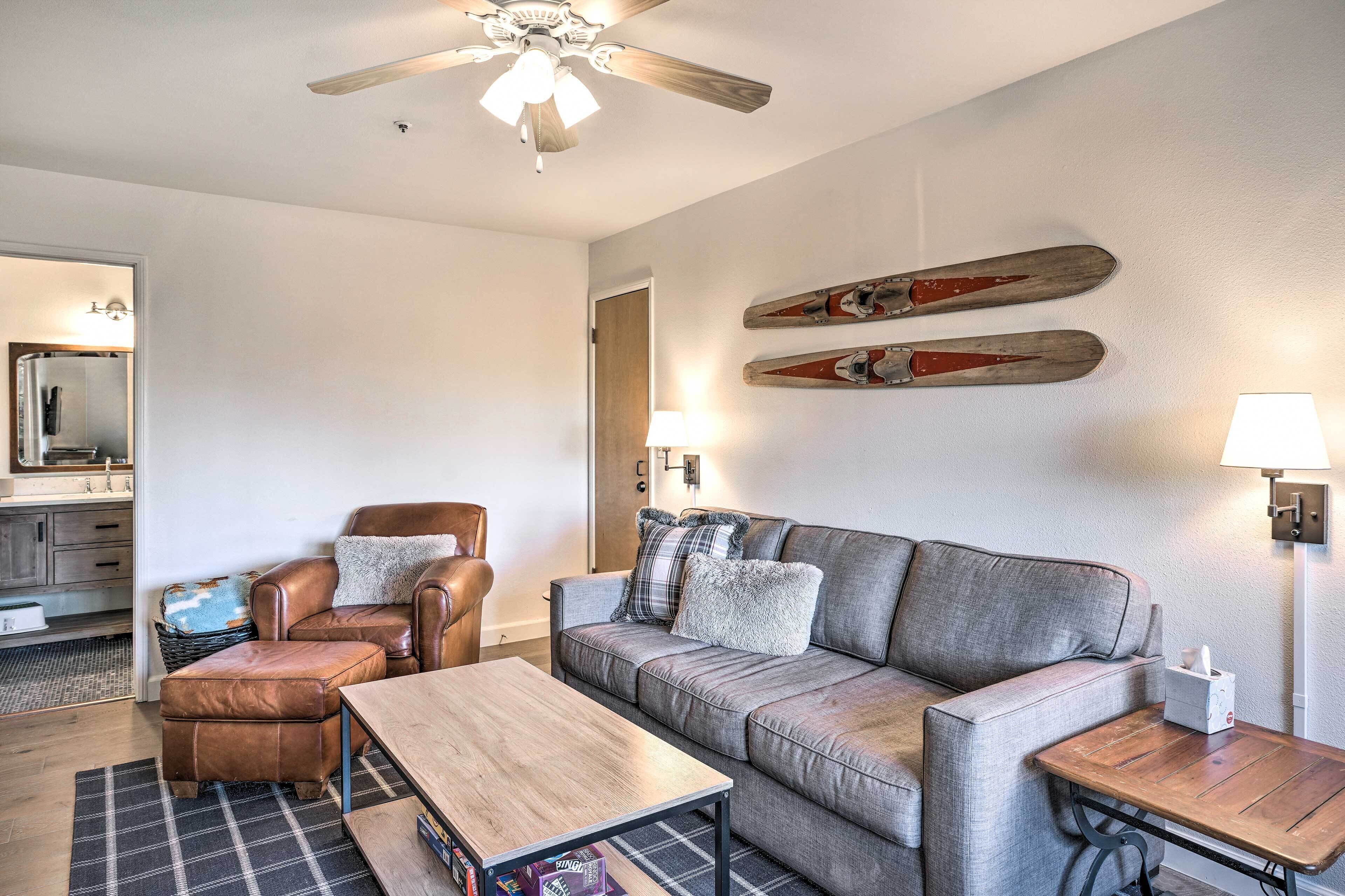 A modern living room with a grey sofa, leather armchair, and wooden coffee table. There are two wall-mounted skis and a ceiling fan.