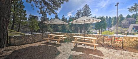 An outdoor patio area with picnic tables and umbrellas, surrounded by stone walls and pine trees.