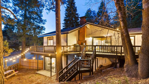 Une cabane à deux étages avec une terrasse, entourée d’arbres, et dotée d’un escalier et de guirlandes lumineuses.
