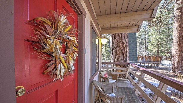 A red door with a corn decoration, a wooden porch with wicker chairs, and a view of a forest.