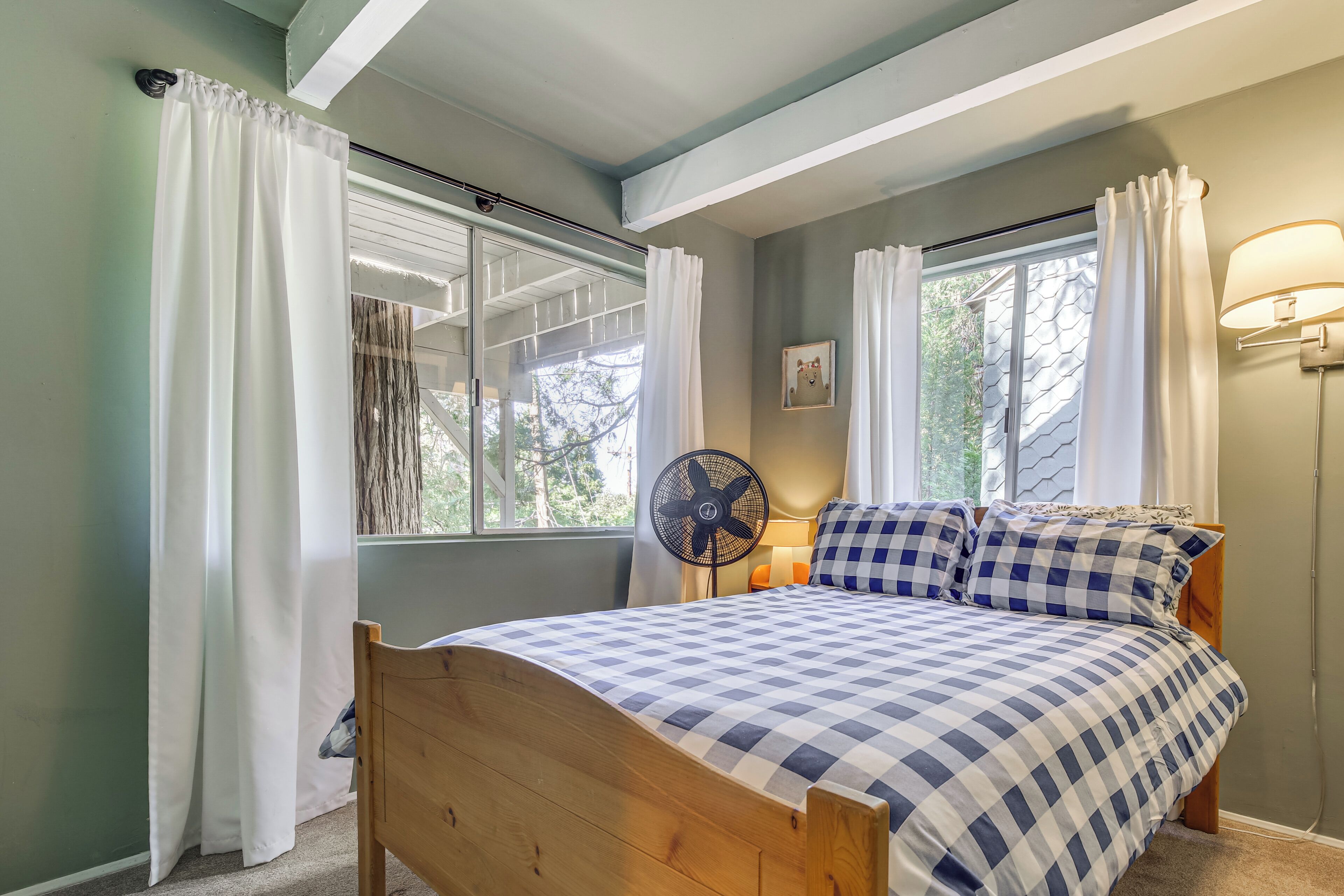 A bedroom with a wooden bed, checkered bedding, a standing fan, and a window with curtains.