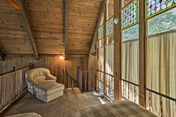 A cozy room with wooden beams, a cushioned chair, a bed, and large windows with curtains. - Stunning Mountain-top Getaway w/ Multi-level Decks (Lake Arrowhead)