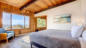 Une chambre dotée d’un grand lit, de poutres apparentes en bois au plafond et de grandes fenêtres donnant sur l’extérieur.