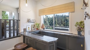 A kitchen with dark cabinetry, a marble countertop, two wooden bar stools, a window with a view of trees, and a hanging light fixture. - Lake Arrowhead Retreat w/ Decks + Mountain Views! (Lake Arrowhead)